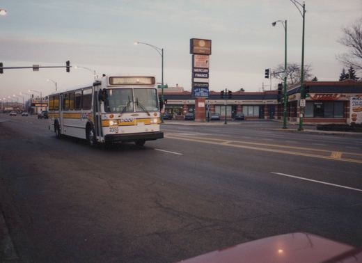A white and yellow Pace bus with number 2355 is driving down a street at dusk with stores and a traffic light in the background.