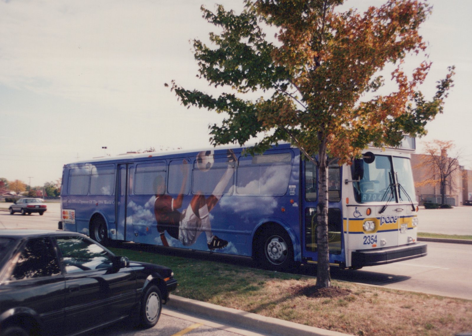 A blue and white Pace motor bus, number 2354, with a soccer player wrap for the 1994 World Cup, is parked on a street with autumn leaves on the trees.