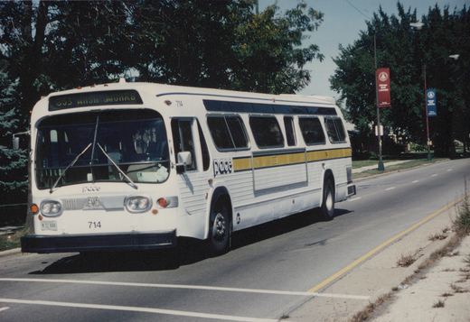A white Pace bus with yellow and blue stripes and the number 714 is driving on a street under a clear sky, with trees and a Chicago State University banner in the background.