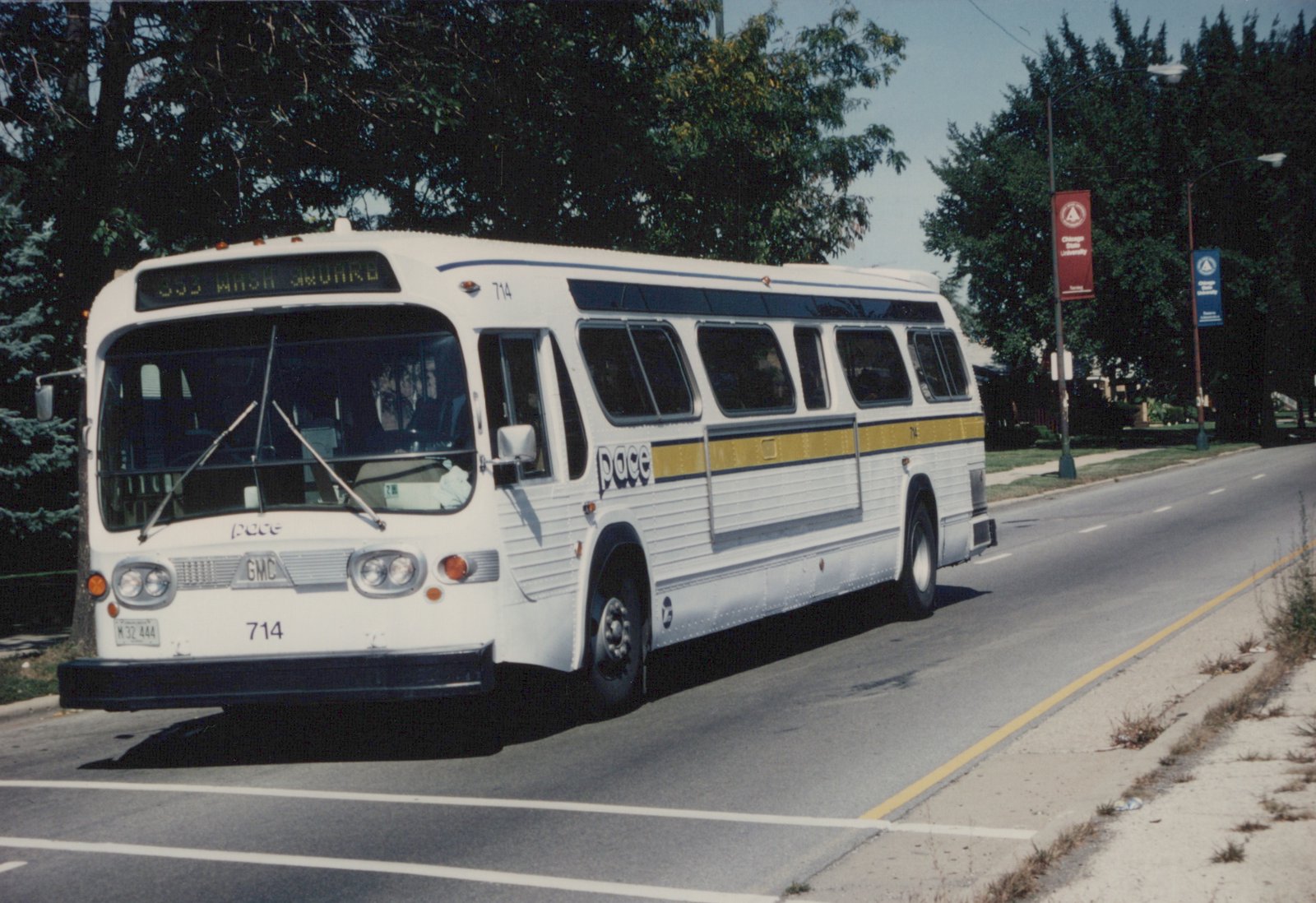 A white Pace bus with yellow and blue stripes and the number 714 is driving on a street under a clear sky, with trees and a Chicago State University banner in the background.