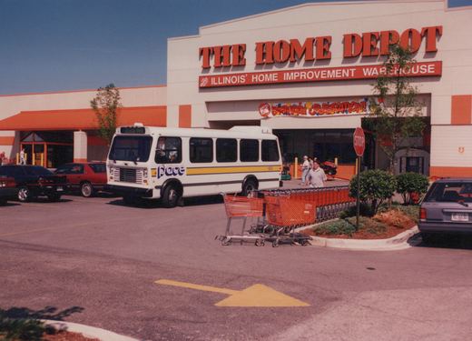 A white Pace bus with yellow and purple stripes is parked in a shopping center lot in front of a Home Depot store. Several cars and shopping carts are visible, along with a few people.