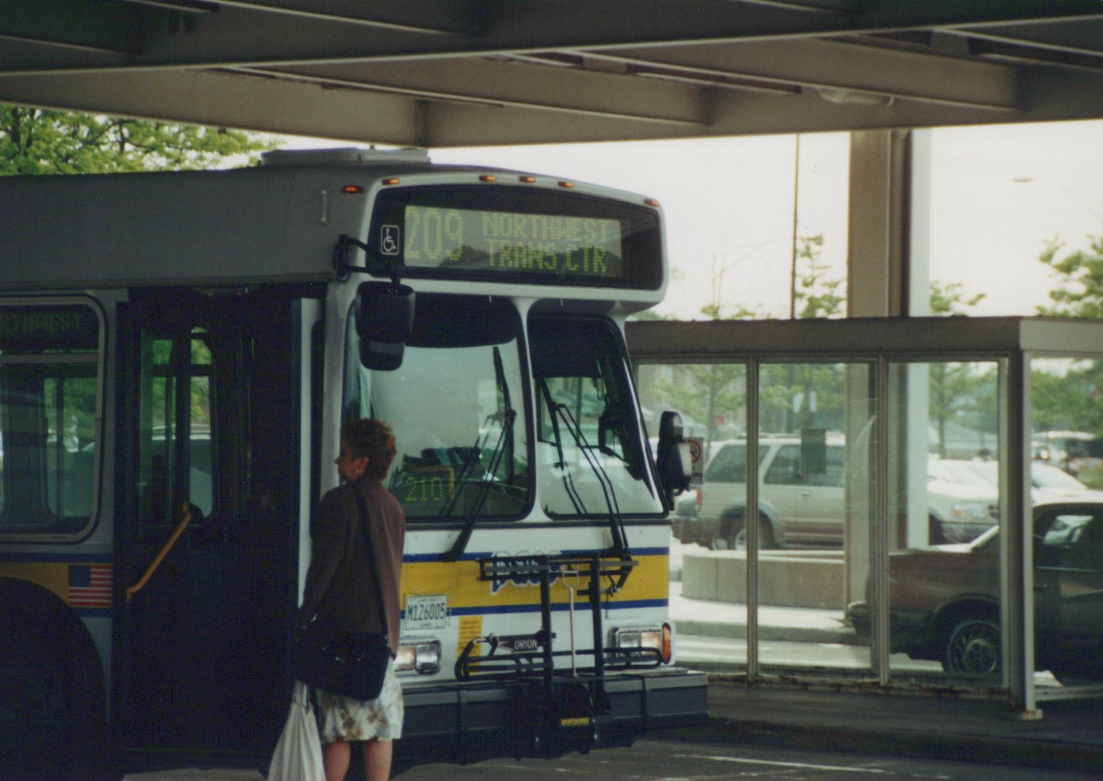 A white and yellow Pace bus with the number 6005 and route 209 to Northwest Trans Ctr is stopped at a sheltered bus terminal with a woman boarding.