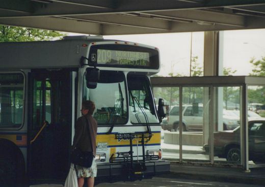 A white and yellow Pace bus with the number 6005 and route 209 to Northwest Trans Ctr is stopped at a sheltered bus terminal with a woman boarding.