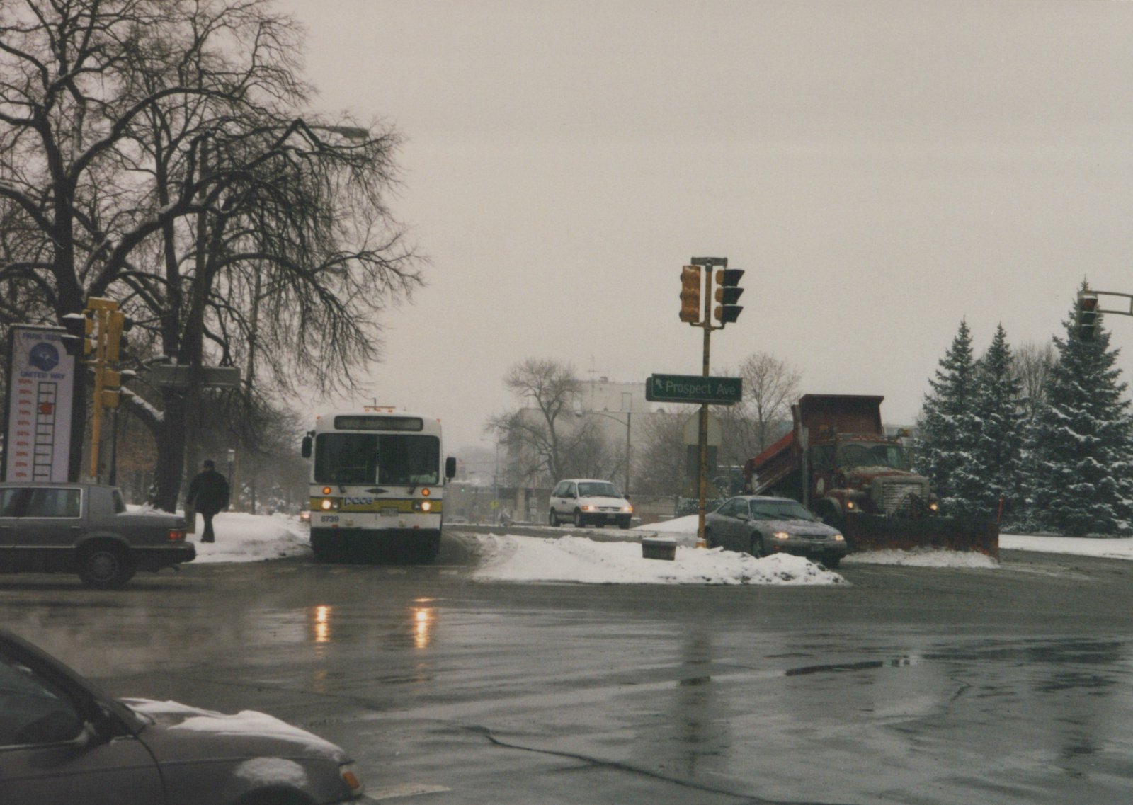 Front view of a white Pace bus numbered 8739 stopped at a snowy street intersection with a green street sign for Prospect Ave and a snowplow truck in the background.