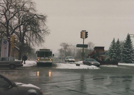 Front view of a white Pace bus numbered 8739 stopped at a snowy street intersection with a green street sign for Prospect Ave and a snowplow truck in the background.