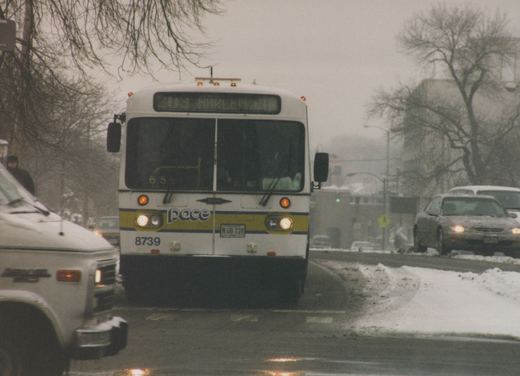 Front view of a white Pace bus with the number 8739 and route 209 Harlem, on a snowy street with cars and bare trees, in daytime.