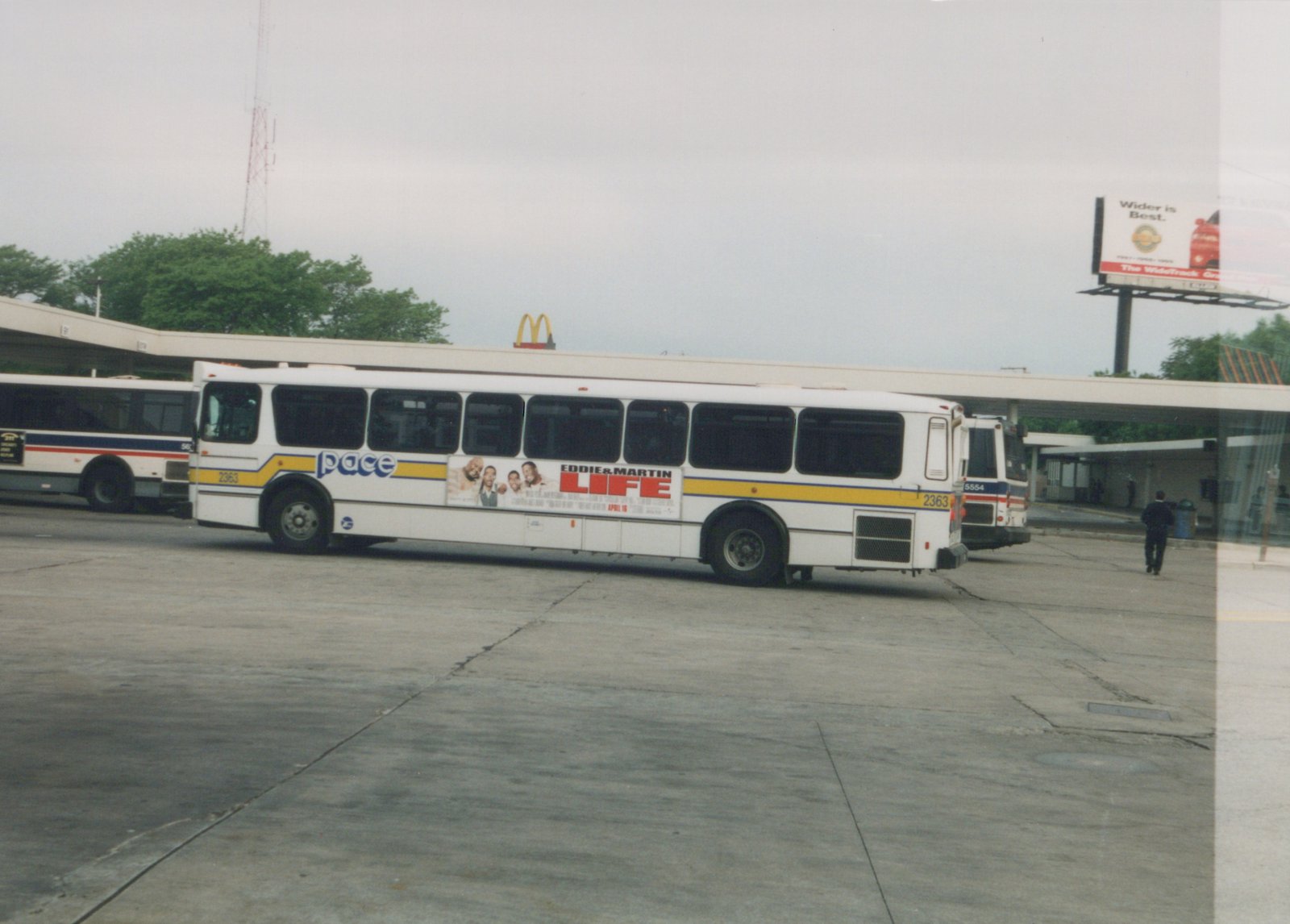 A white Pace bus with the number 2363 is parked at a bus terminal, with a movie advertisement on its side and other buses in the background.