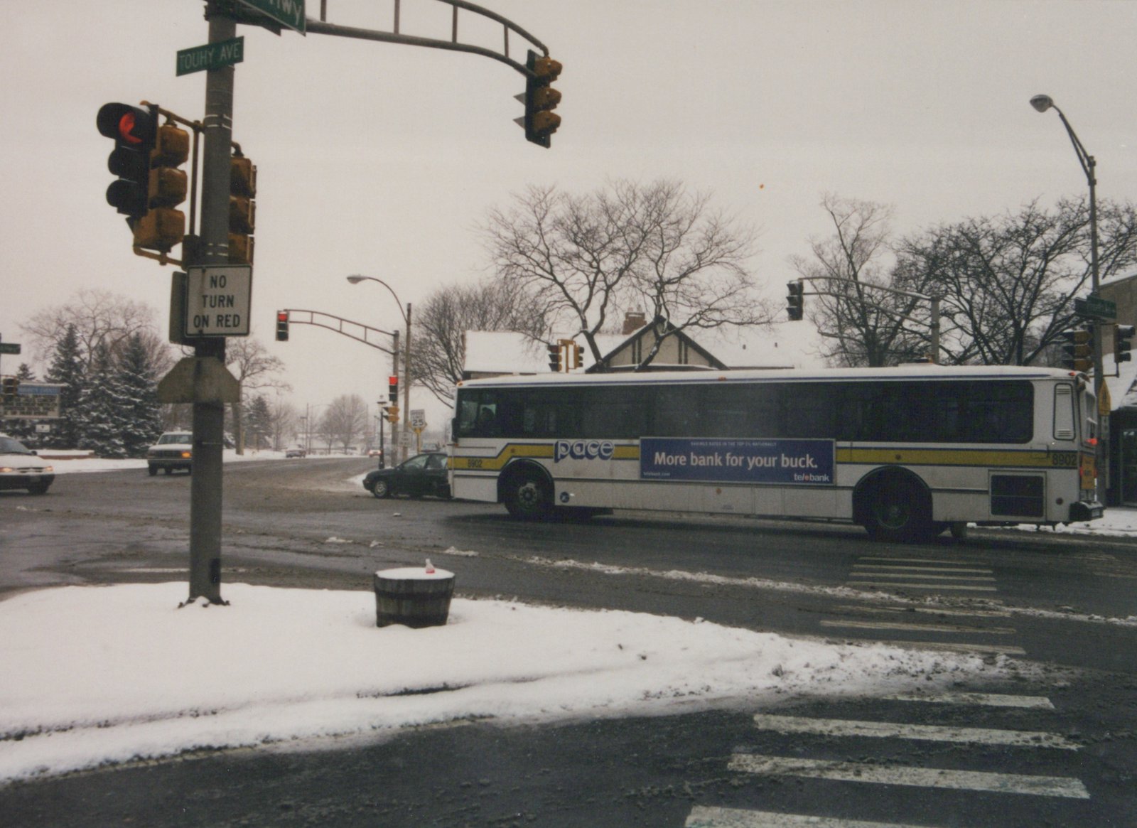 A white Pace motor bus with the number 8902 is stopped at a snow-dusted street corner in a suburban setting, with street signs for Touhy Avenue and a 'No Turn on Red' sign visible.