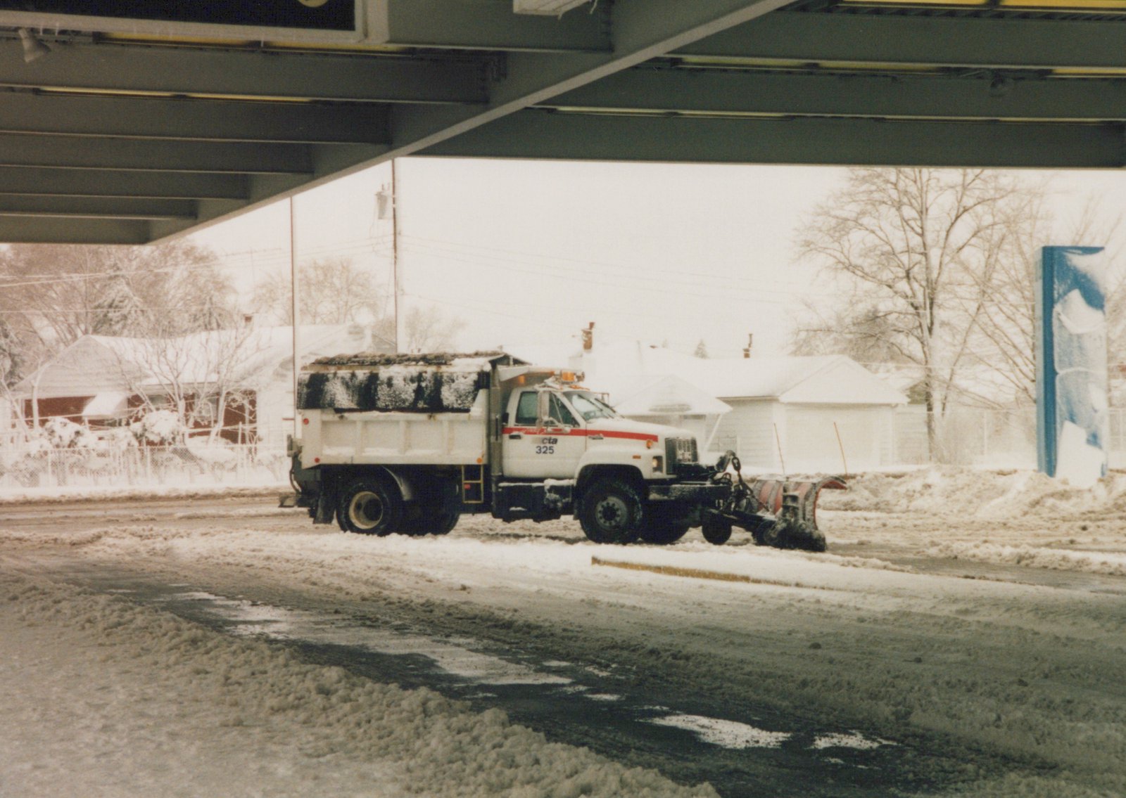A white CTA snowplow truck with the number 325 is plowing a snowy street under a metal overpass, with snow-covered houses in the background.