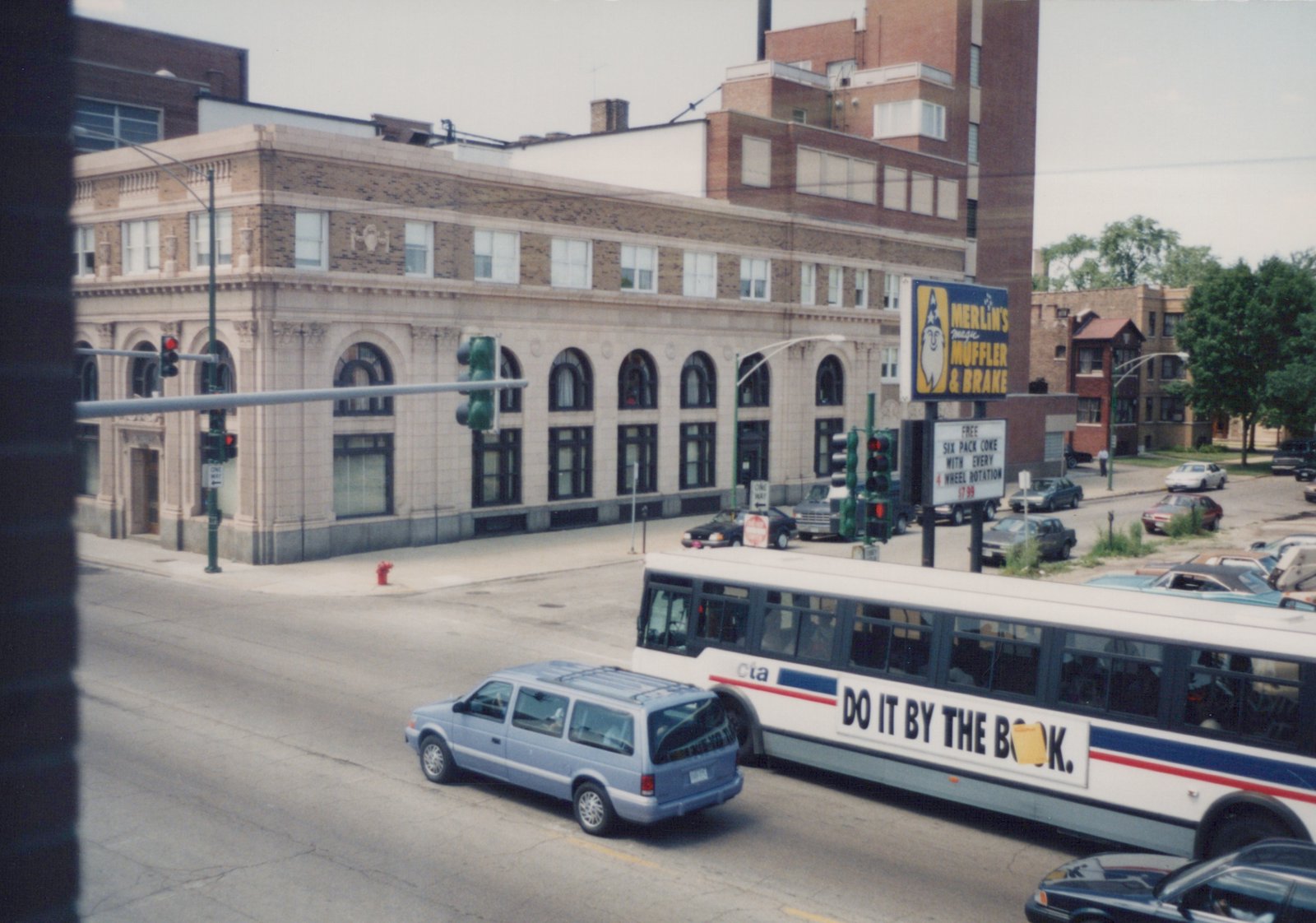 A white CTA bus with blue and red stripes drives down a city street next to a light blue minivan, with a building and a Merlin's Muffler & Brake sign in the background.