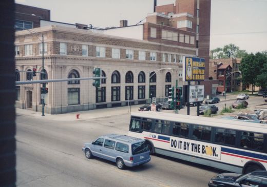 A white CTA bus with blue and red stripes drives down a city street next to a light blue minivan, with a building and a Merlin's Muffler & Brake sign in the background.