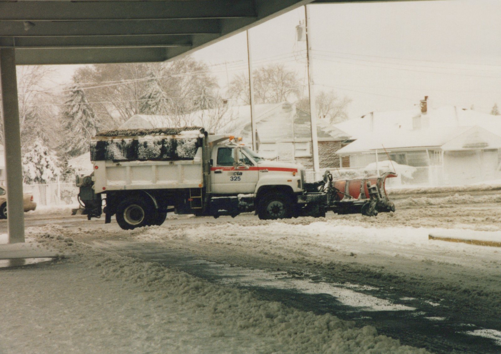 A white CTA snowplow truck with number 325 is clearing a street covered in snow and slush, with snow-covered trees and houses in the background and an elevated structure overhead.