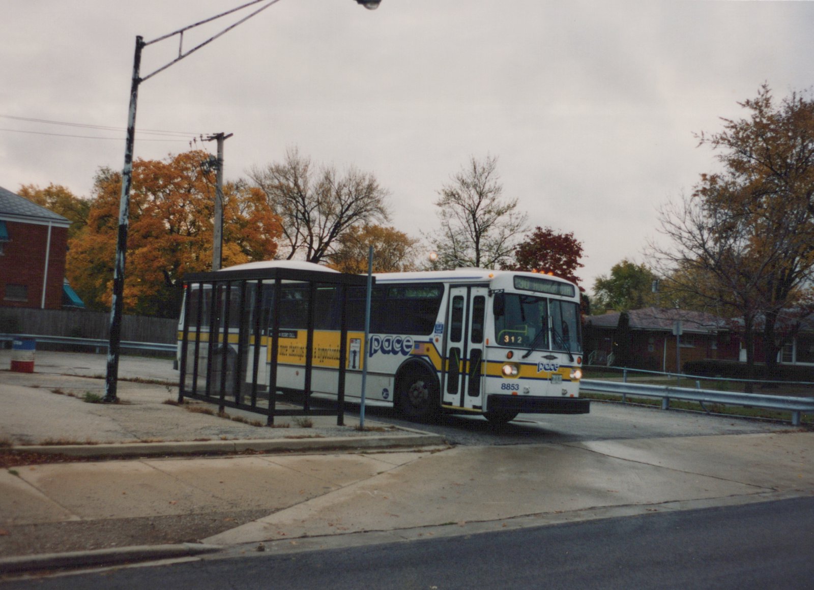 A white and yellow Pace bus, number 8853, is parked at a bus stop on a street with autumn trees in the background.