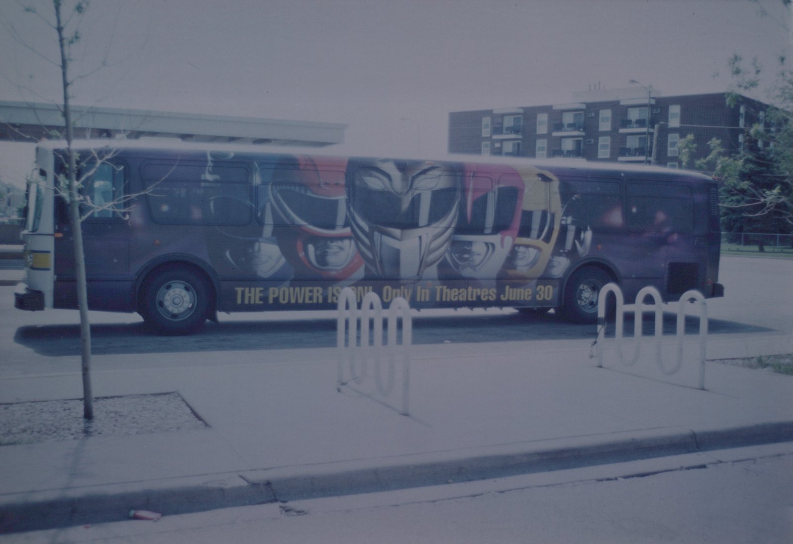 A purple CTA bus with a Power Rangers movie ad on its side, parked at a curb next to a bike rack and a young tree, with an apartment building in the background.