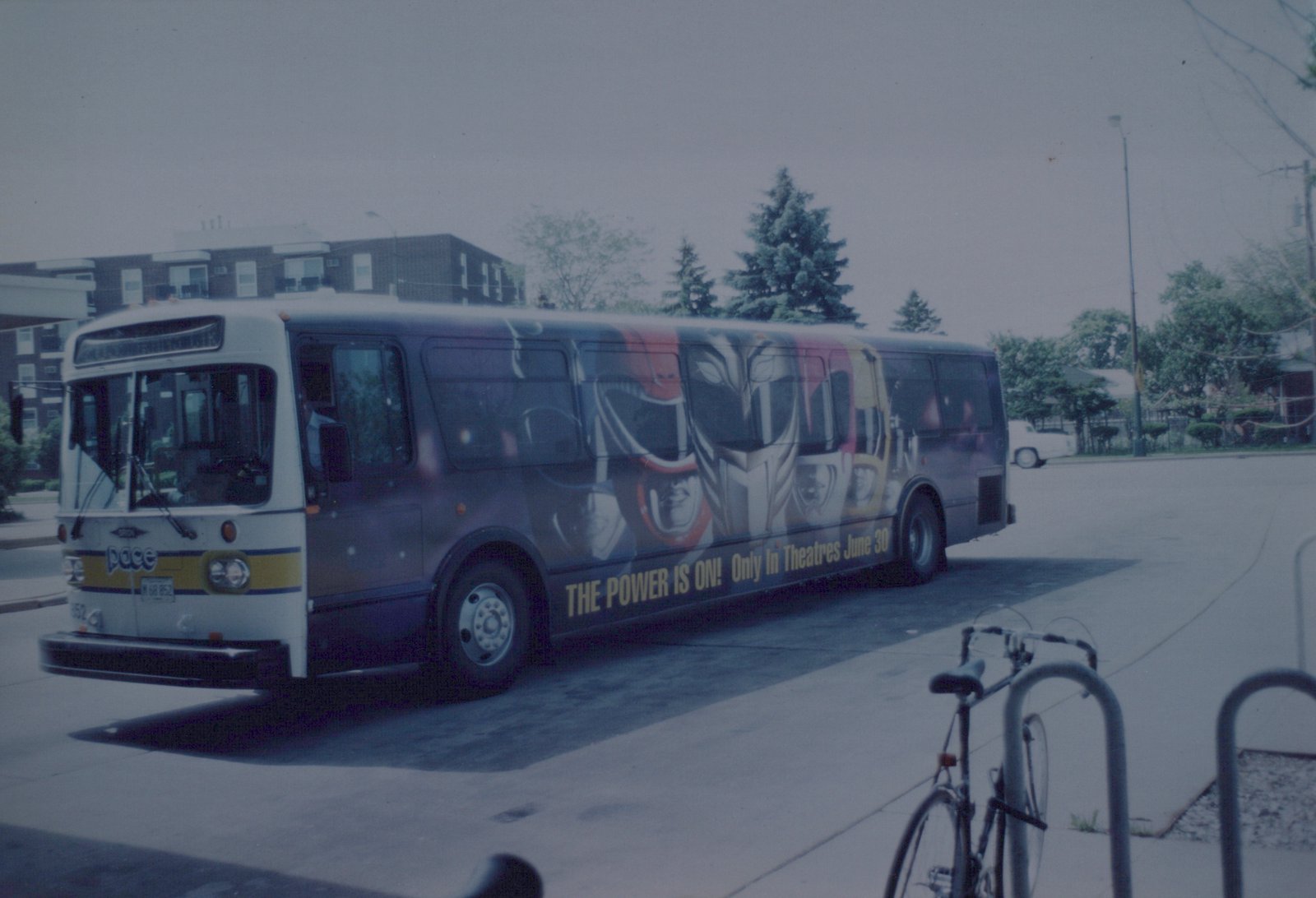 A white and blue Pace motor bus, number 8852, is parked on a street with a 'Power Rangers' movie ad on its side. A bicycle is locked to a rack nearby.