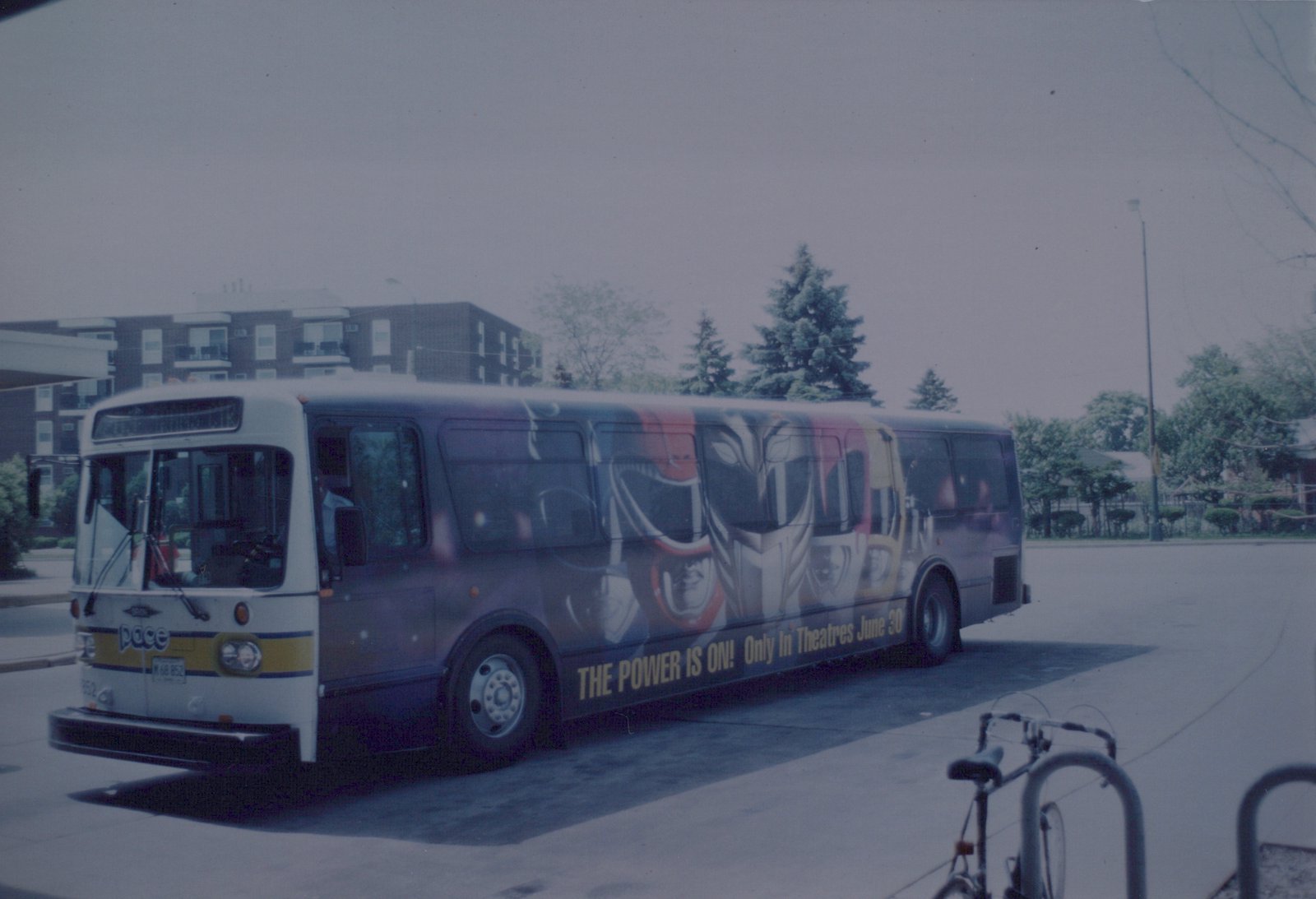 A white and yellow Pace motor bus with a large Power Rangers movie advertisement on its side is parked on a street. A bicycle is locked to a rack in the foreground.