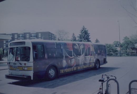 A white and yellow Pace motor bus with a large Power Rangers movie advertisement on its side is parked on a street. A bicycle is locked to a rack in the foreground.