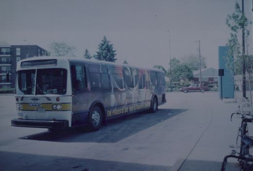 A white and yellow Pace bus with the number 8852 is parked on a street corner, with a full-wrap advertisement for a movie on its side. A maroon car is visible in the background, and the scene is set under an overcast sky.