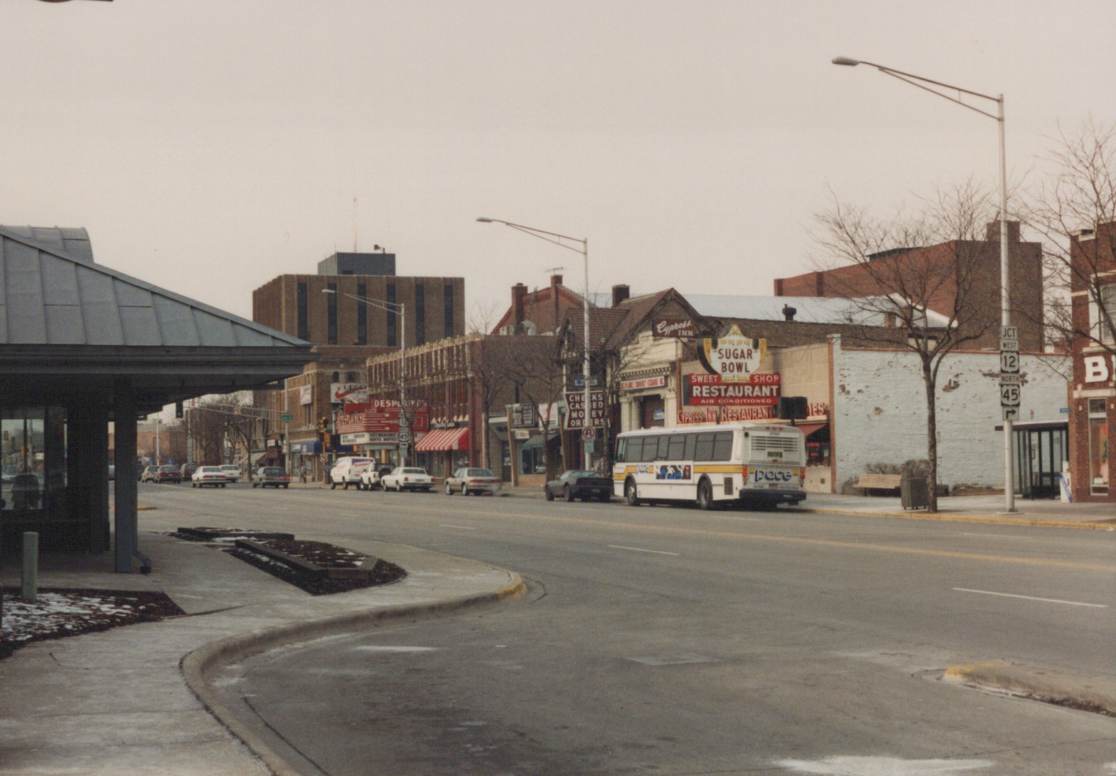 A white Pace motor bus with the number 8799 is parked on a city street lined with brick buildings and storefronts, including a Sugar Bowl restaurant, on a cloudy winter day.