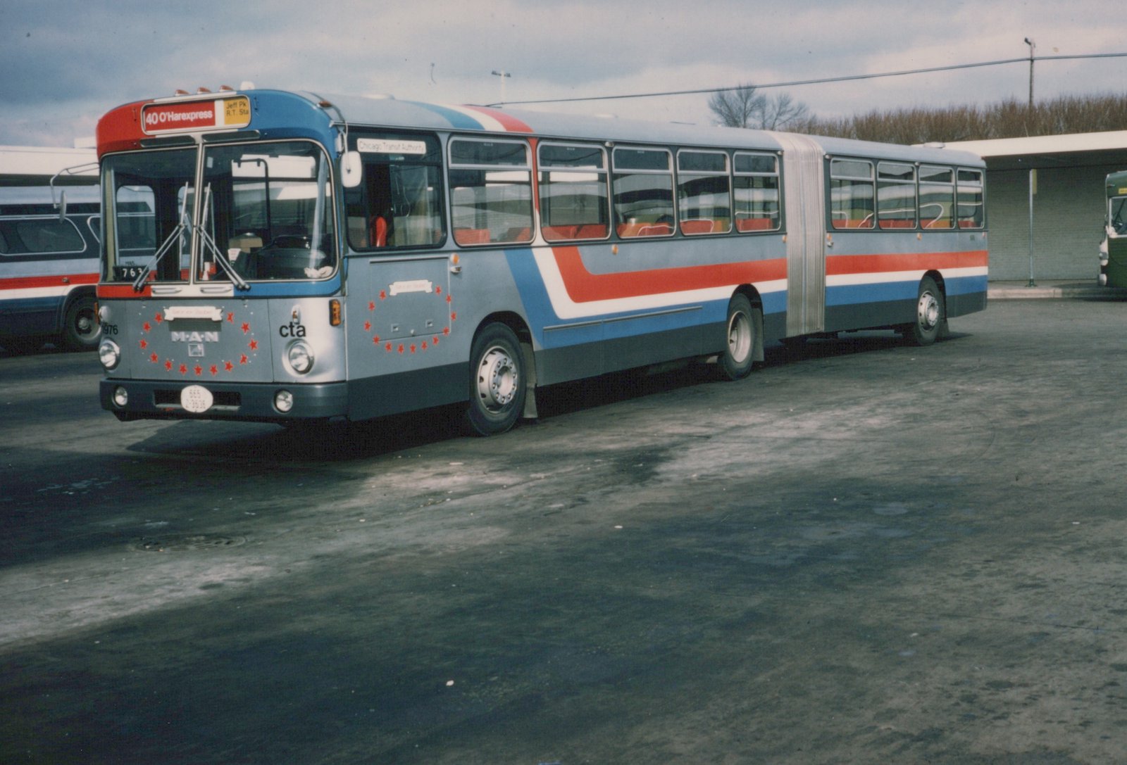 A silver articulated bus with red and blue stripes and red stars, labeled CTA and number 1976, parked in a paved yard under an overcast sky.