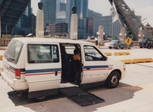 A white paratransit van with blue and red stripes and the lettering CDT is parked on a bridge with its side door open and a ramp extended to the sidewalk. A person in a wheelchair is visible inside. The background shows a city skyline and other vehicles on the bridge.