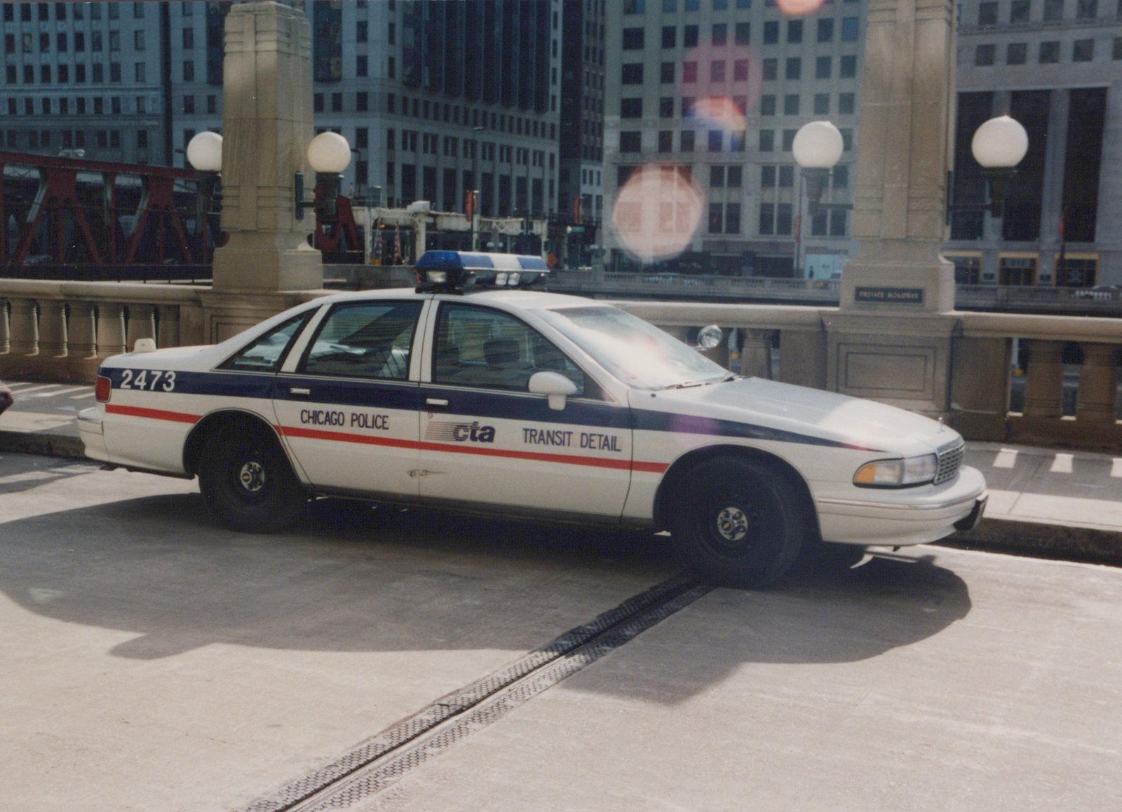 A white and blue Chicago Police CTA Transit Detail car, number 2473, is parked on a city bridge on a sunny day with tall buildings in the background.