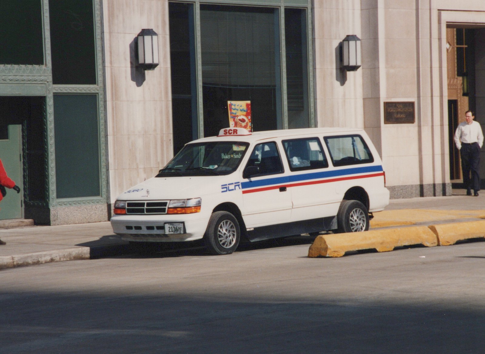 A white van with blue and red stripes and 'SCR' lettering is parked on a sunny city street, with pedestrians walking on the sidewalk.