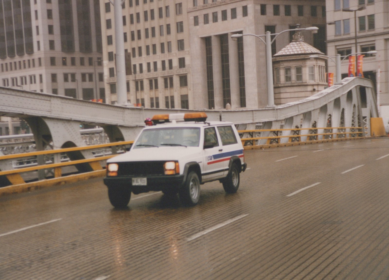 A white Chicago Transit Authority utility SUV with blue and red stripes drives on a wet bridge with a large building and a bridge operator's house in the background.