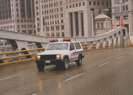 A white Chicago Transit Authority utility SUV with blue and red stripes drives on a wet bridge with a large building and a bridge operator's house in the background.