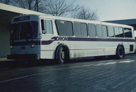 A white Orion I motor bus with the number 1750 displayed on the front and side, featuring a blue and red stripe design, is parked on a street with bare trees in the background.