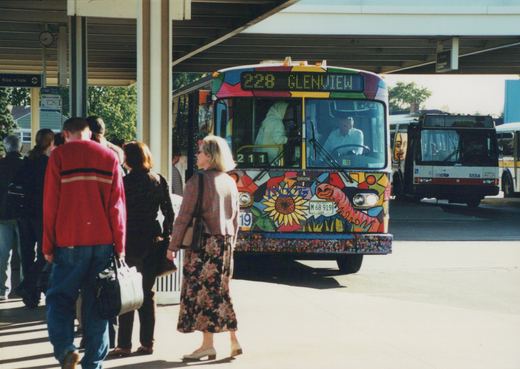 A colorful Pace bus with a mosaic design and the number 211 is stopped at an outdoor terminal, with people walking nearby and another bus visible in the background.