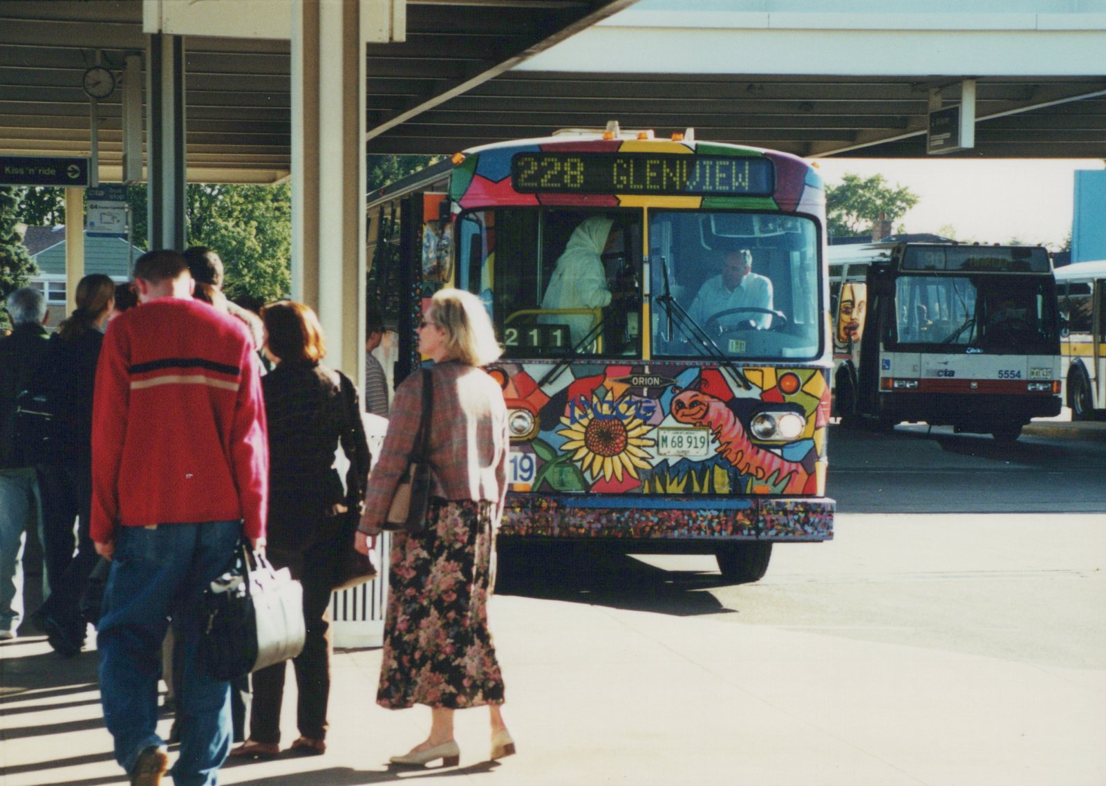 A colorful Pace bus with a mosaic design and the number 211 is stopped at an outdoor terminal, with people walking nearby and another bus visible in the background.