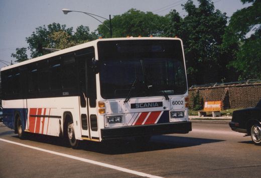 A white, red, and blue CTA bus with the number 6002 on its front, driving down a street with trees and a brick wall in the background.
