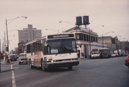 A white and orange CTA bus with '22 TO HARRISON' on its sign is driving on a city street with a water tower and brick buildings in the background.