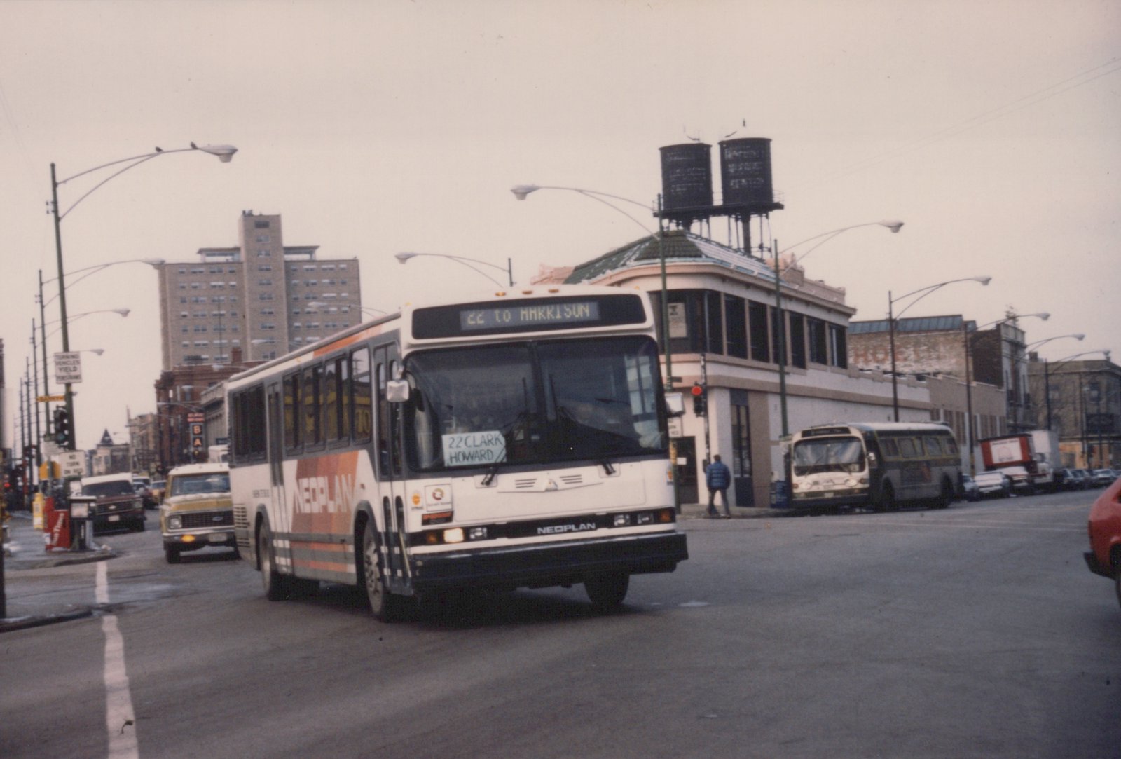 A white and orange CTA bus with '22 TO HARRISON' on its sign is driving on a city street with a water tower and brick buildings in the background.