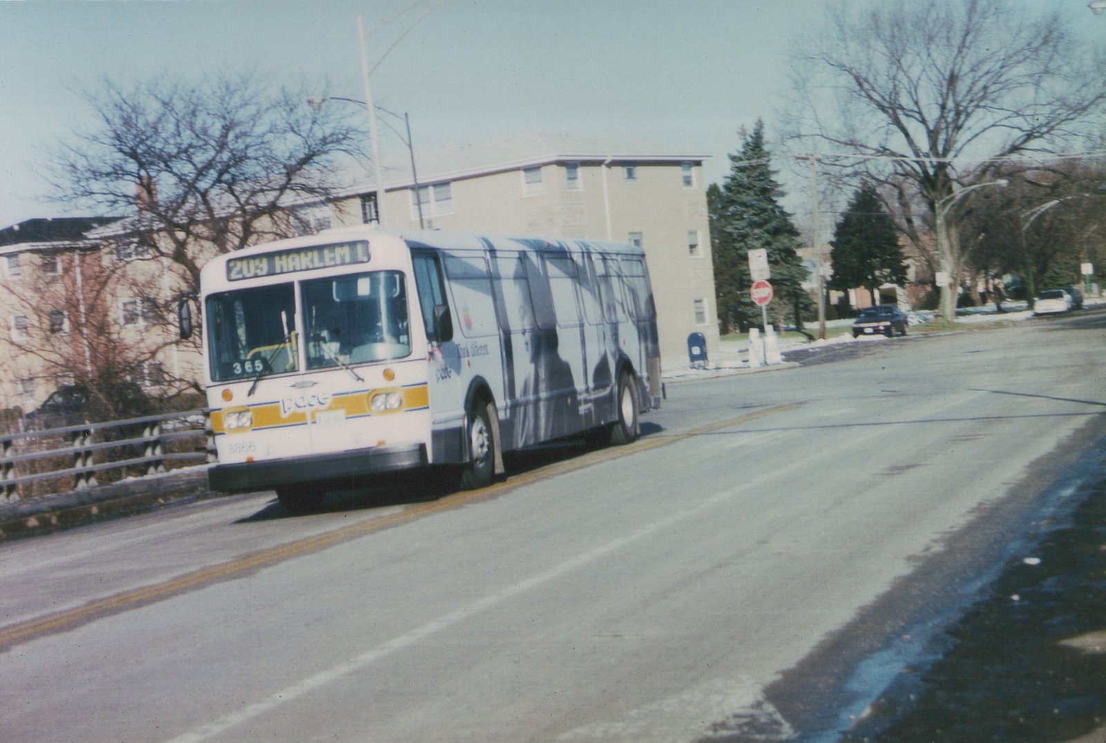 A white Pace bus with a yellow stripe and a large gray Apple 'Think different' ad on its side is driving down a tree-lined street with snow on the ground.