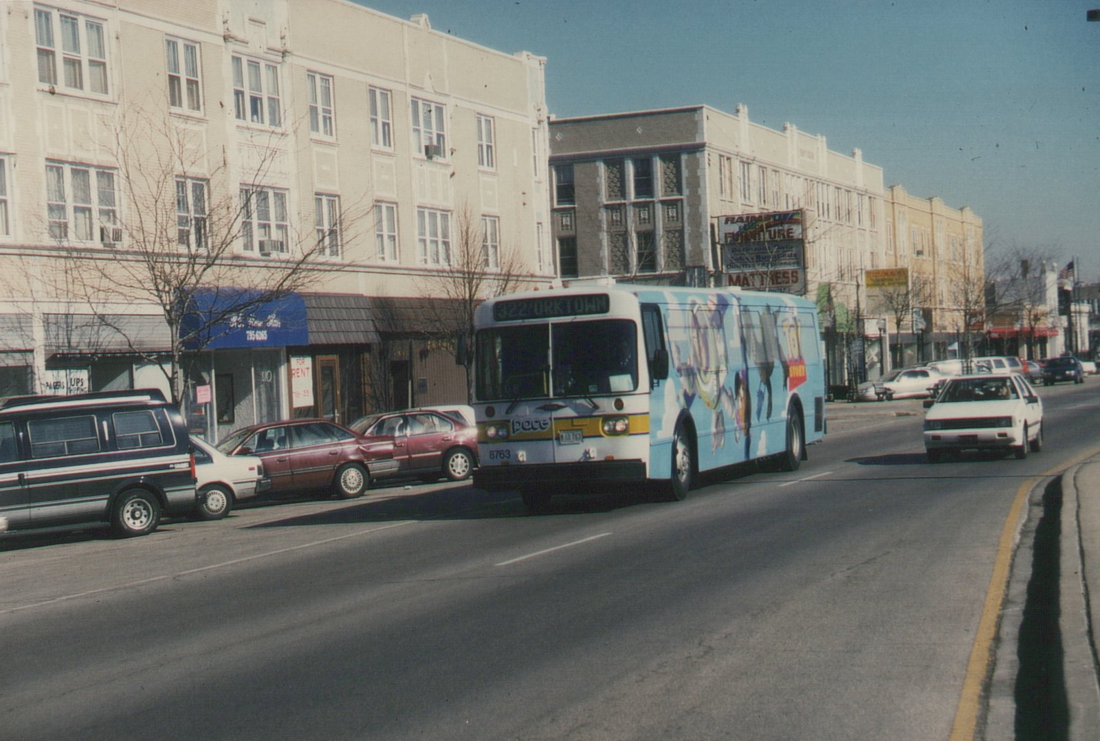 A white and blue Pace bus with a Toy Story ad drives down a city street lined with parked cars and storefronts on a sunny day.