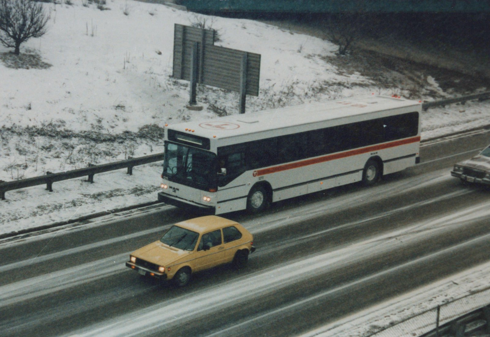 A white and orange RTA bus with the number 1211 is driving on a snow-covered highway, with a yellow hatchback car nearby.