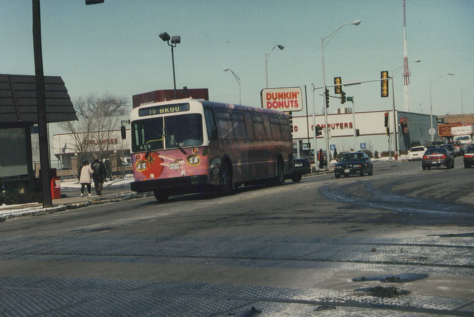 A white and red Pace bus with the number 609 on its front is driving down a wet, icy street in a commercial area during winter, with people walking on the sidewalk and a Dunkin' Donuts sign in the background.