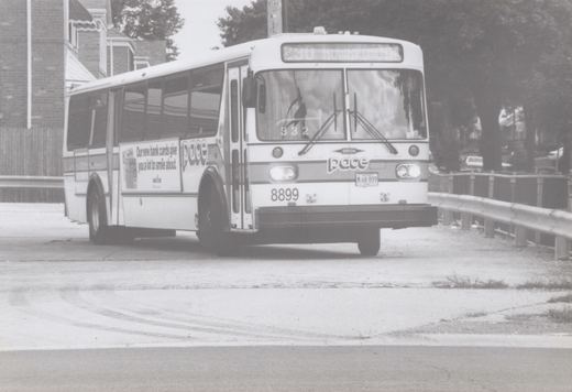 A front three-quarter view of a white Pace bus with the number 8899, parked on a street with a guardrail and trees in the background.