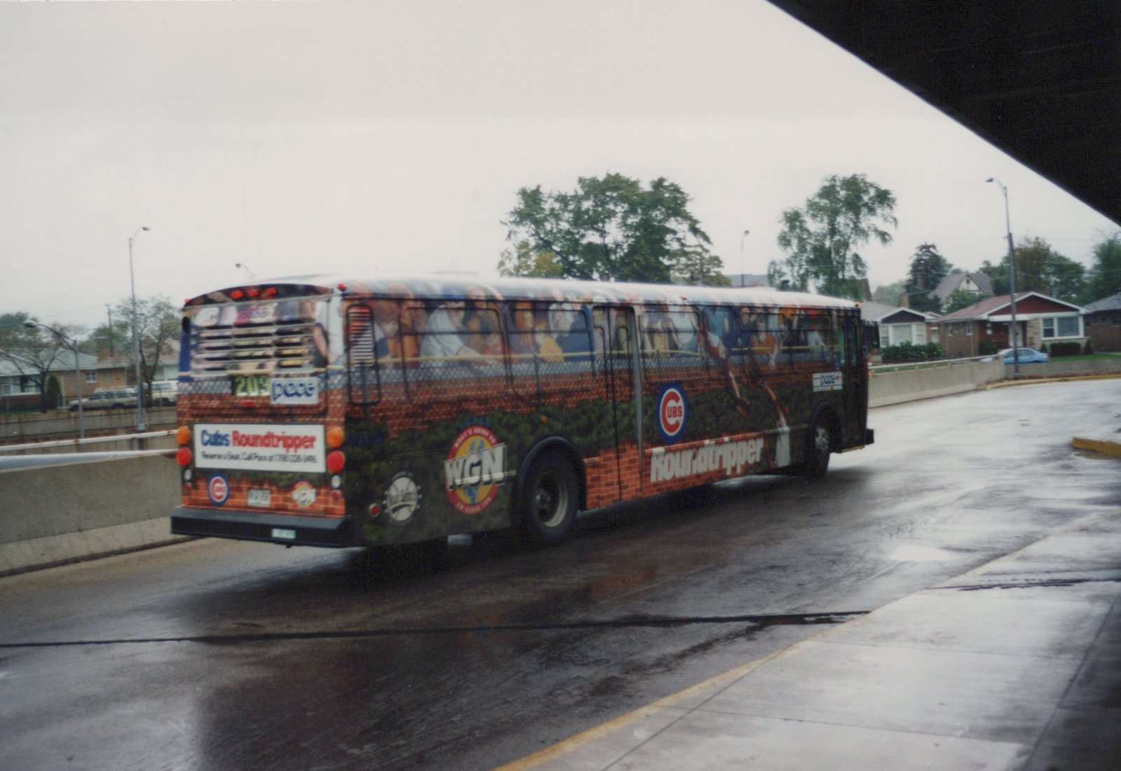 A large, colorful Pace bus with a Chicago Cubs advertisement is driving on a wet road under a concrete overpass on a cloudy day.