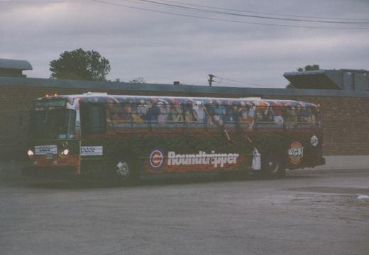 A black Pace bus with a full wrap advertisement for the Chicago Cubs 'Roundtripper' is parked on a paved lot. The livery features fans, a brick wall, and ivy. The Pace logo is visible on the front and side, and the number 8858 is on the front. A WGN logo is visible on the rear.