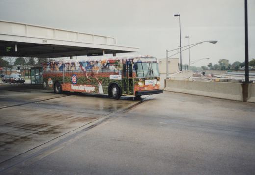 A single Pace motor bus with a Chicago Cubs advertisement is stopped at a modern bus terminal on a cloudy day. The wet pavement reflects the scene, and the bus's fleet number 8858 is visible on the front bumper.