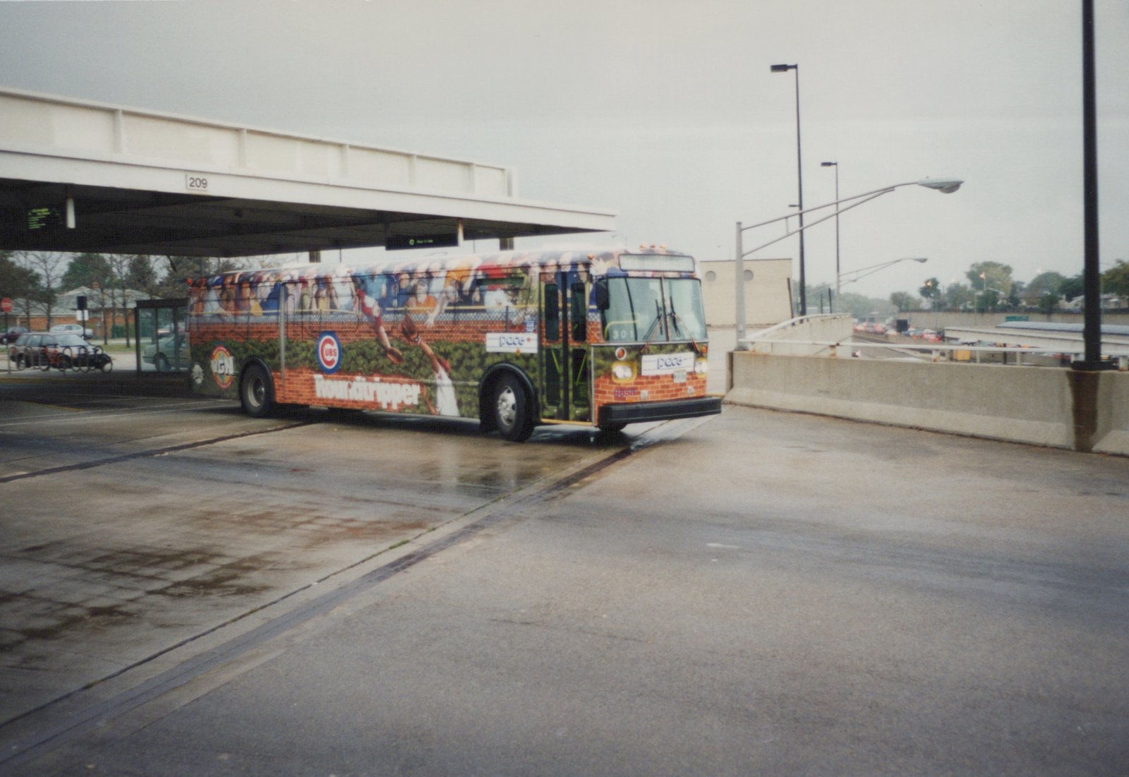 A single Pace motor bus with a Chicago Cubs advertisement is stopped at a modern bus terminal on a cloudy day. The wet pavement reflects the scene, and the bus's fleet number 8858 is visible on the front bumper.