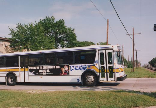 A white Pace bus with yellow and blue stripes, fleet number 8872, featuring a side ad for The X-Files, is parked on a street with trees and a clear sky.