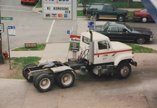A white CTA bus chassis, numbered 217, is parked on a city street with cars and a gas station in the background.