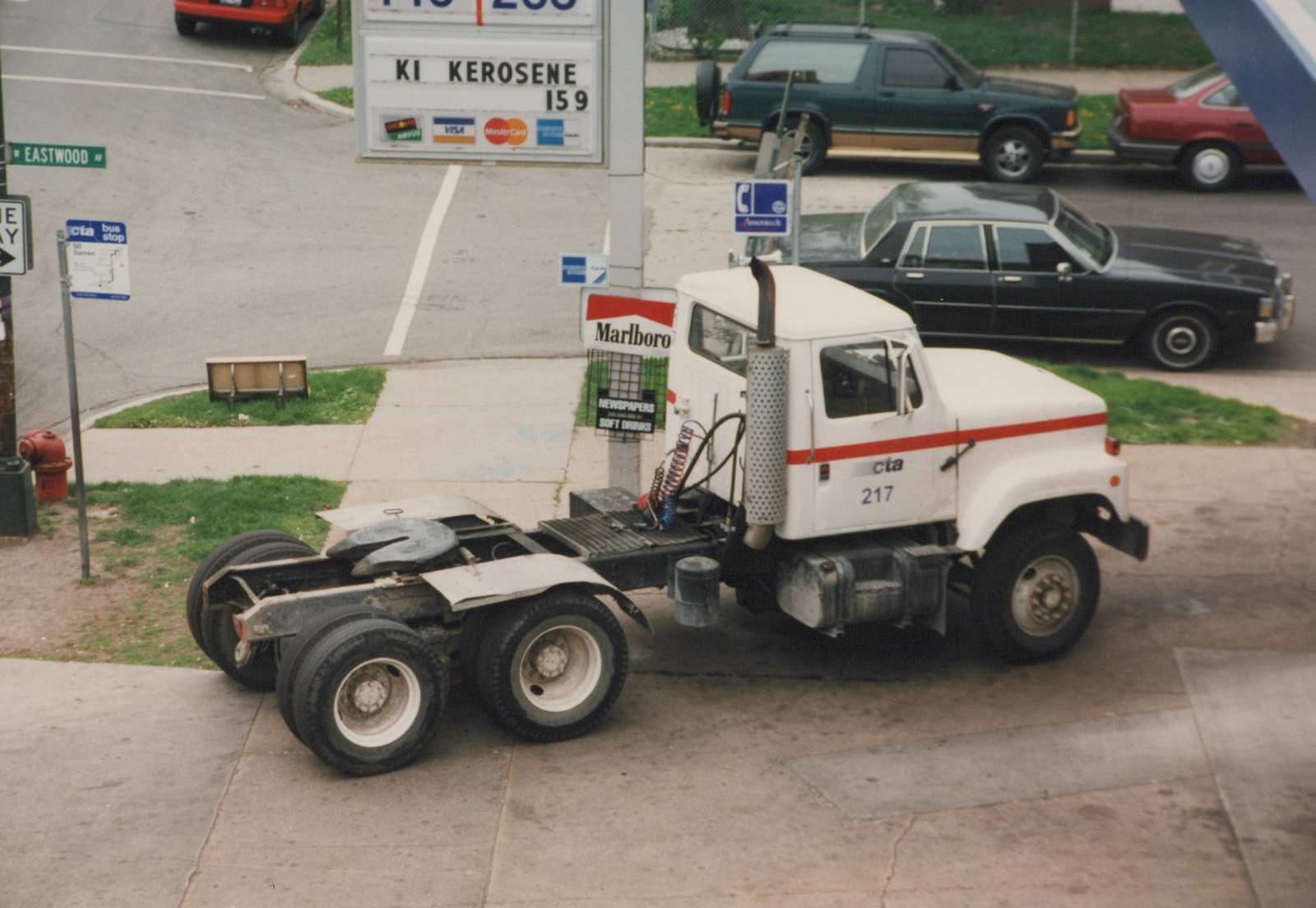 A white CTA bus chassis, numbered 217, is parked on a city street with cars and a gas station in the background.