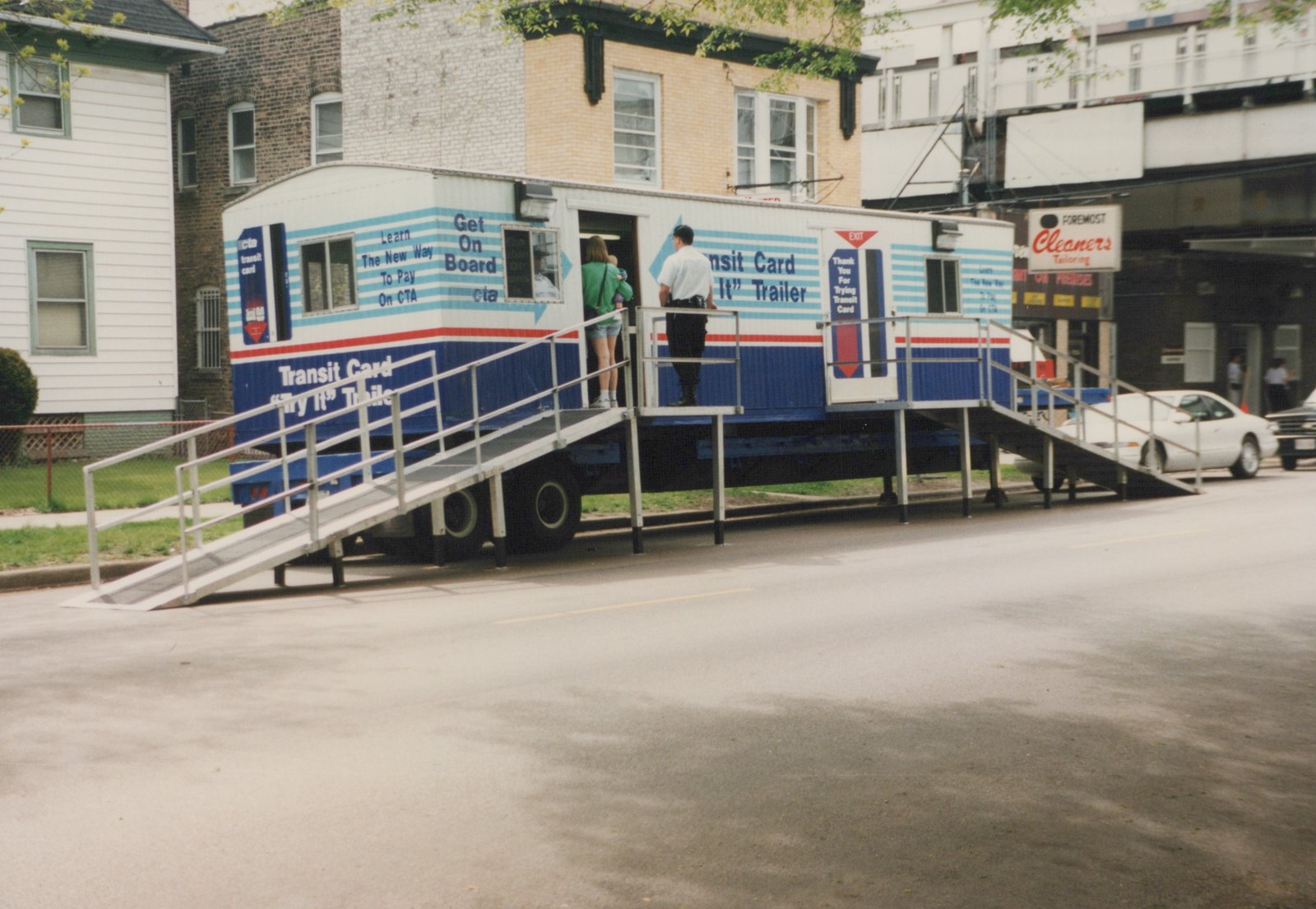 A blue and white CTA trailer with ramps for public access, featuring text about the Transit Card, parked on a street with buildings and cars in the background.