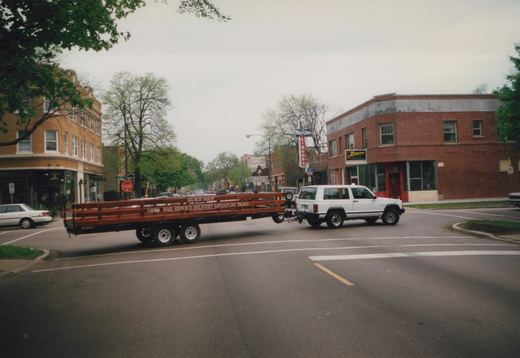 A white SUV tows a long, brown flatbed trailer with 'cta' and 'Skokie Shop' lettering down a street lined with storefronts, including a liquor store and a Zenith sign, under an overcast sky.