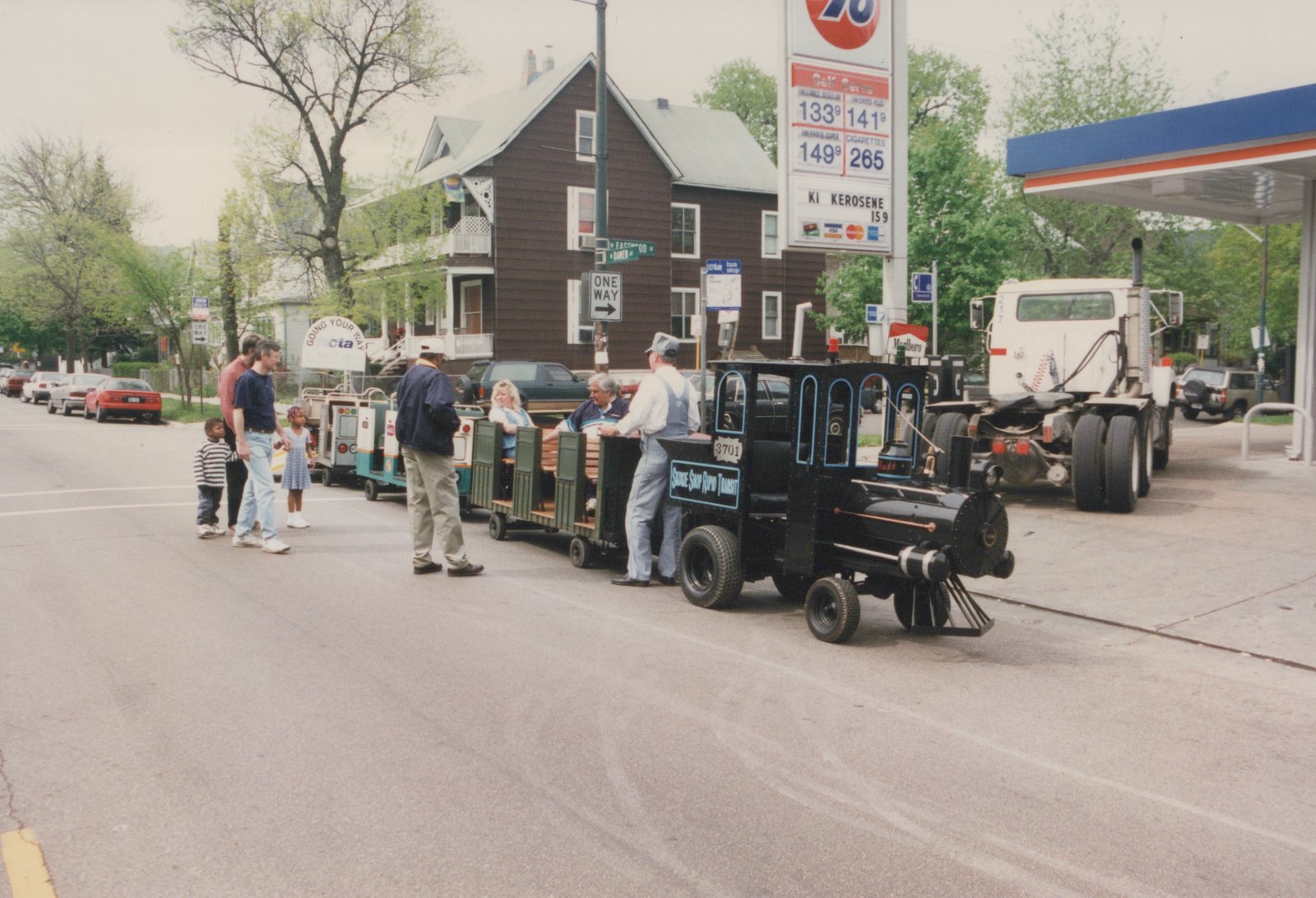 A black, toy-like train labeled 'Smokie Shop Rapid Transit' with number 3701 is on a city street, carrying passengers, with people standing nearby and a gas station in the background.